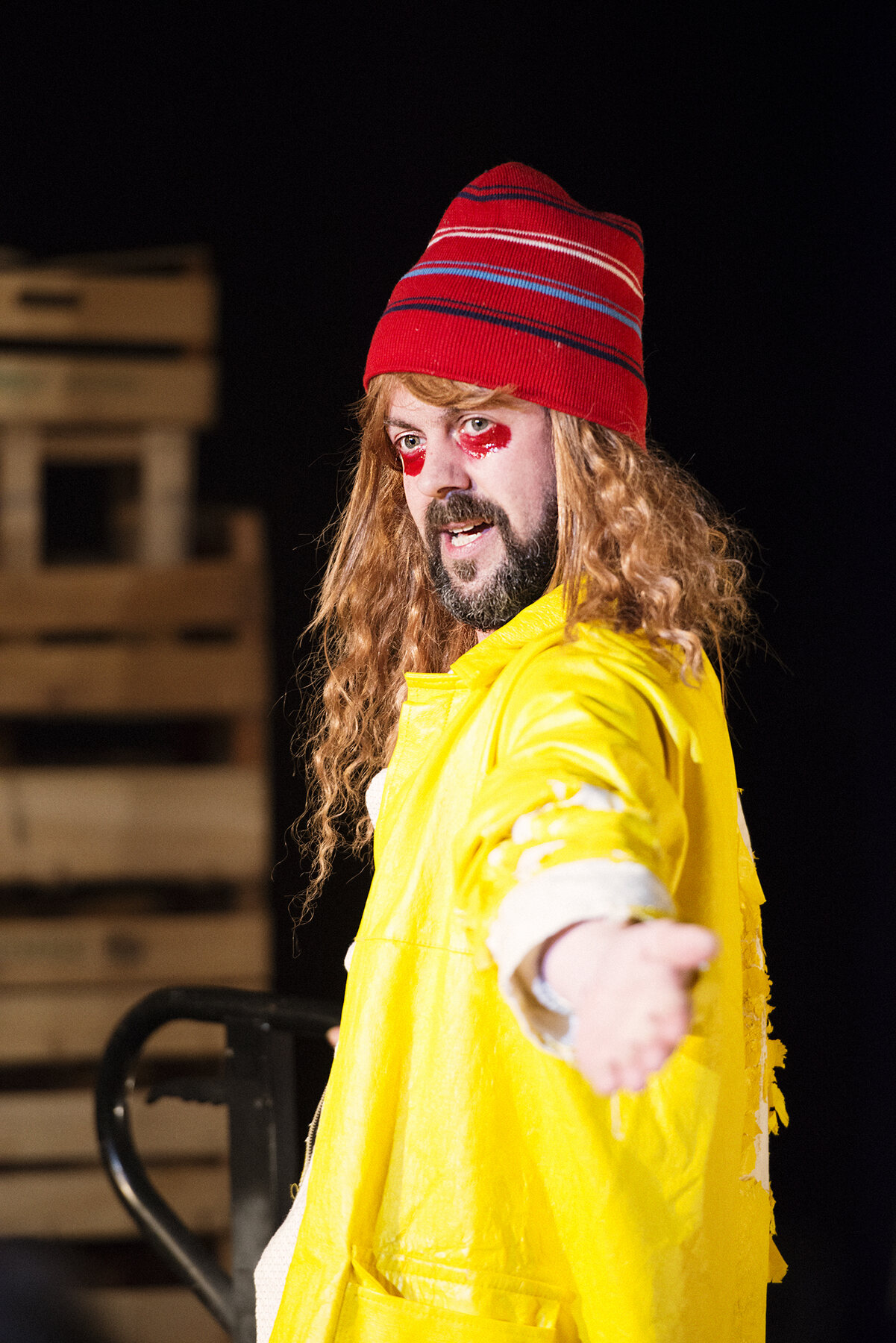 Ein erschöpfter Clown mit roter Mütze und gelbem Mantel streckt eine Hand aus.