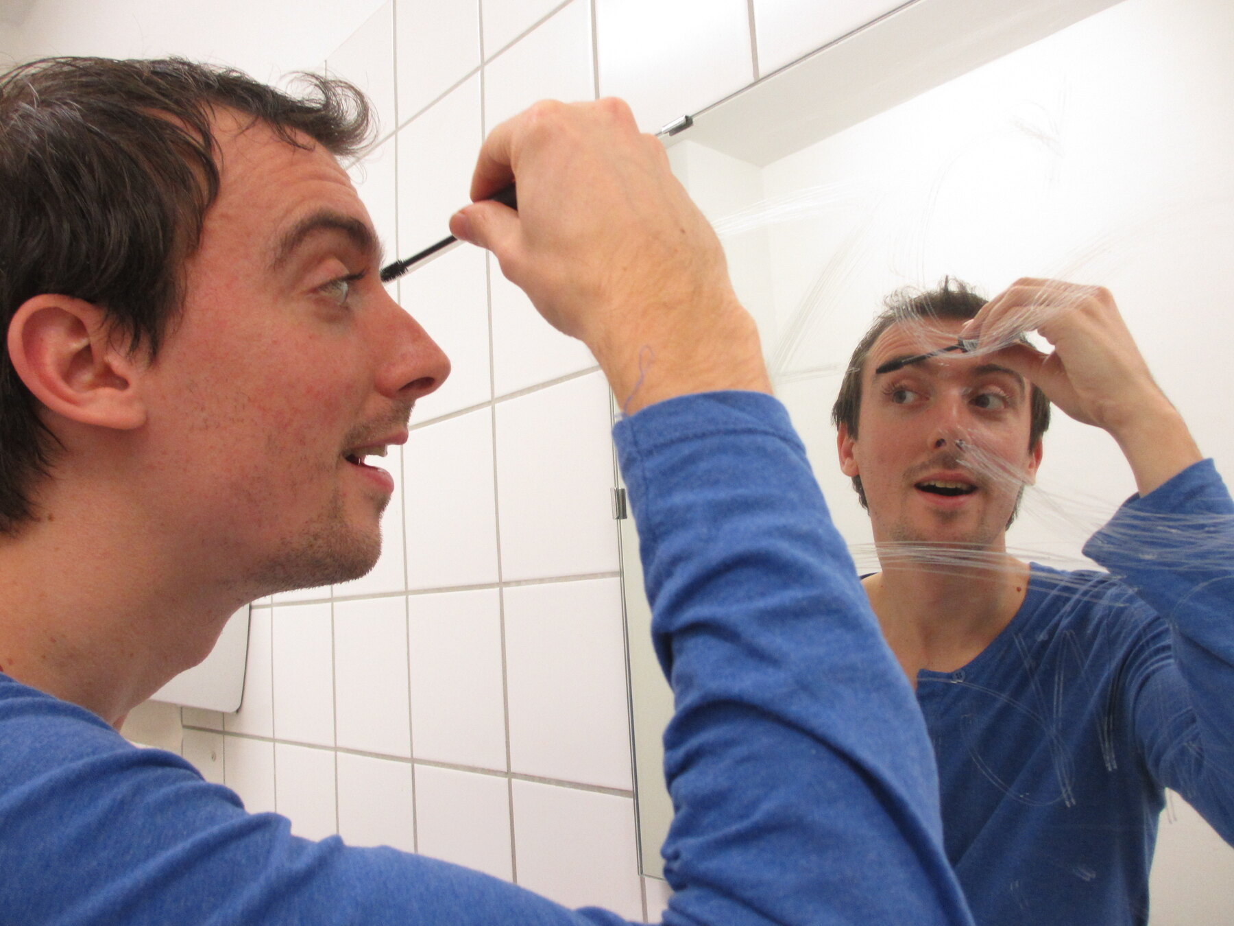 Ein Darsteller schminkt sich vor einem Spiegel in einem Badezimmer.